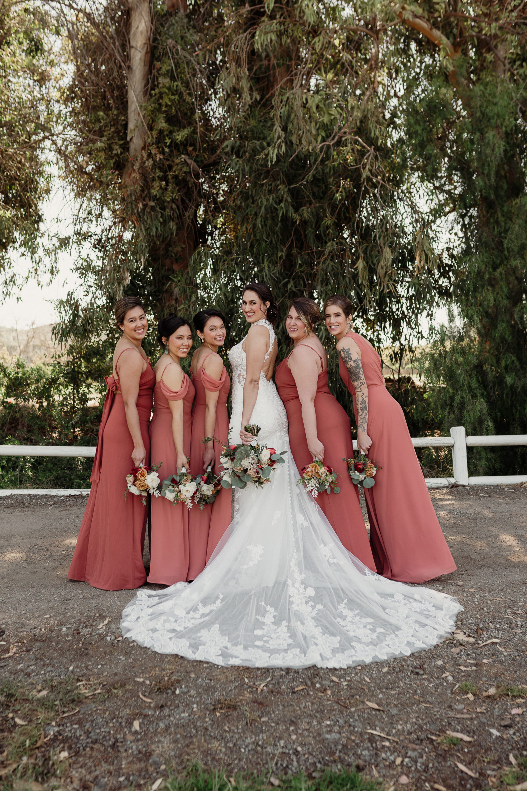 bride wearing a white gown and bridesmaids wearing terracotta dresses hold white rose and eucalyptus bouquets and pose in a line on the road in front of tall eucalyptus trees at walnut grove weddings in moorpark