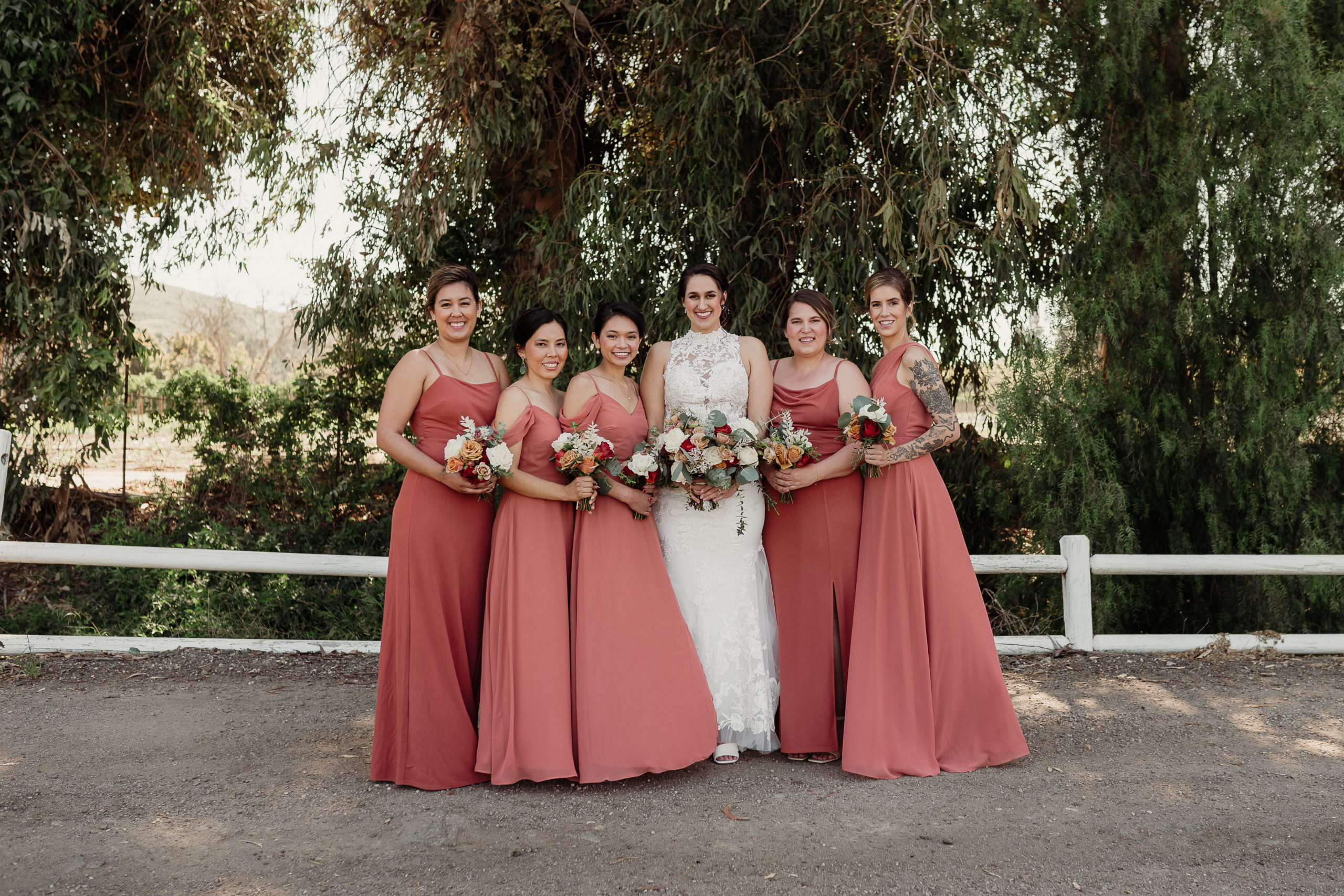 bride wearing a white gown and bridesmaids wearing terracotta dresses hold white rose and eucalyptus bouquets and pose in a line on the road in front of tall eucalyptus trees at walnut grove weddings in moorpark