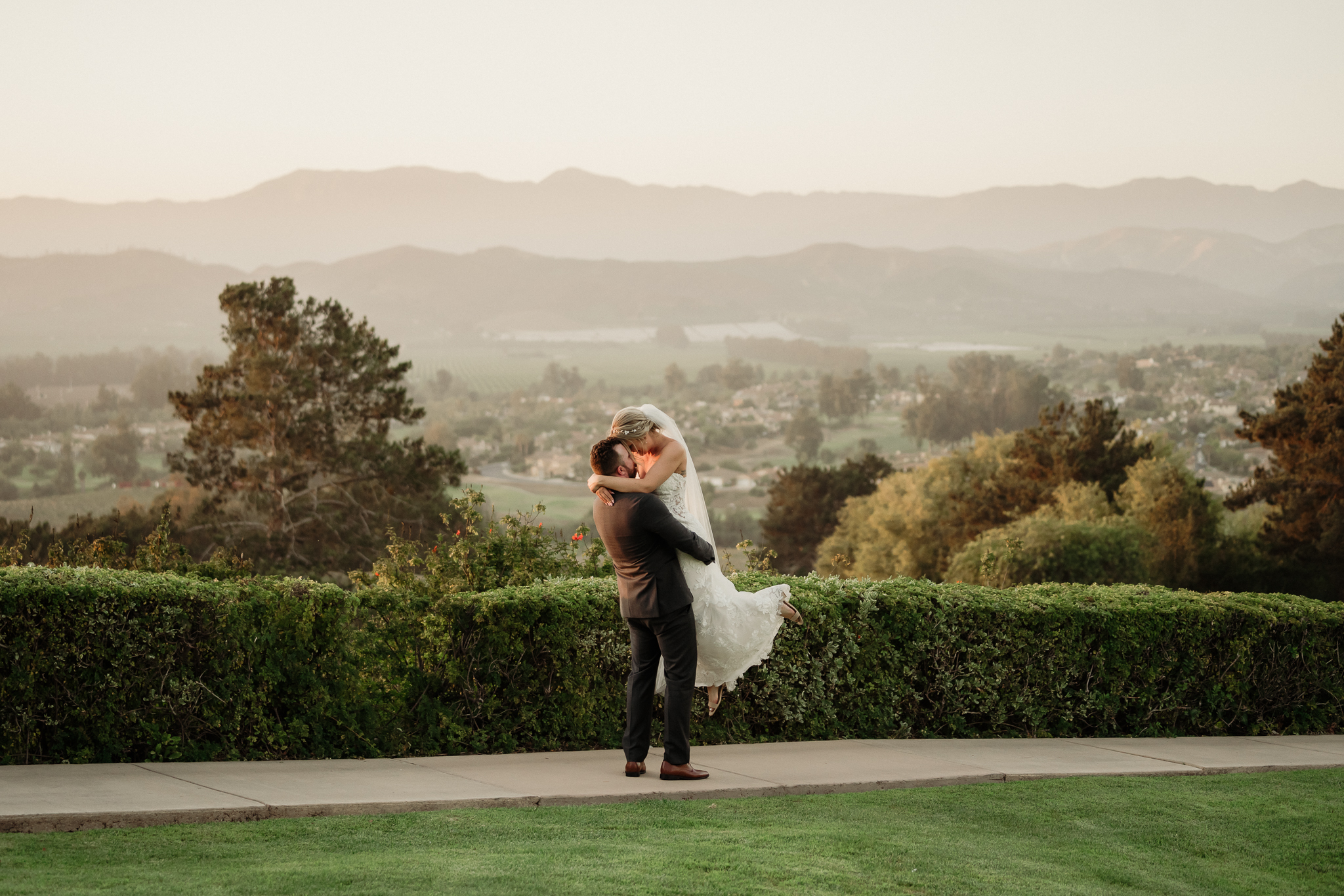 groom picks up his bride for a romantic kiss on top of a mountain overlooking the entire city of Camarillo, CA at Spanish Hill Country Club