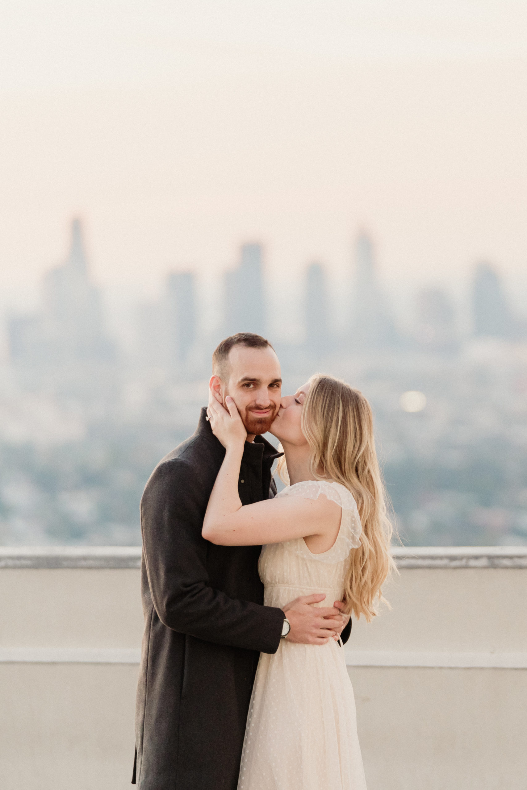 woman wearing an ivory dress kissing her fiance on the cheek at their griffith observatory engagement session overlooking the Los Angeles skyline at sunrise