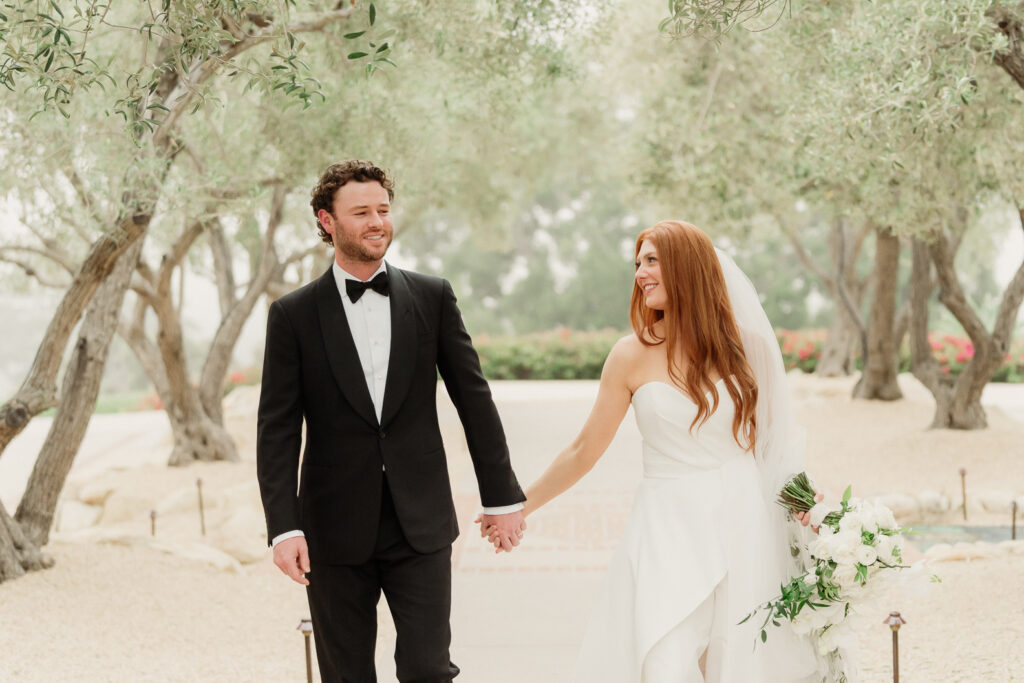 bride with red hair and a white bouquet and groom wearing a traditional black and white tux walk holding hands through the olive trees at the Montecito Club in Santa Barbara, Montecito, CA captured by award winningSanta Barbara wedding photographer Magnolia West Photography