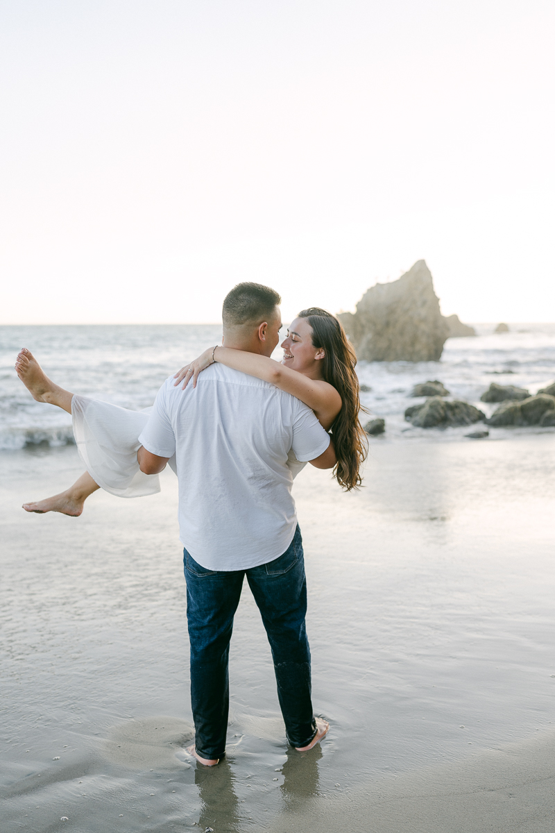 engaged couple man picks up his fiance and carries her on the beach at sunset at el matador beach during their engagement session captured by malibu wedding photographer magnolia west photography