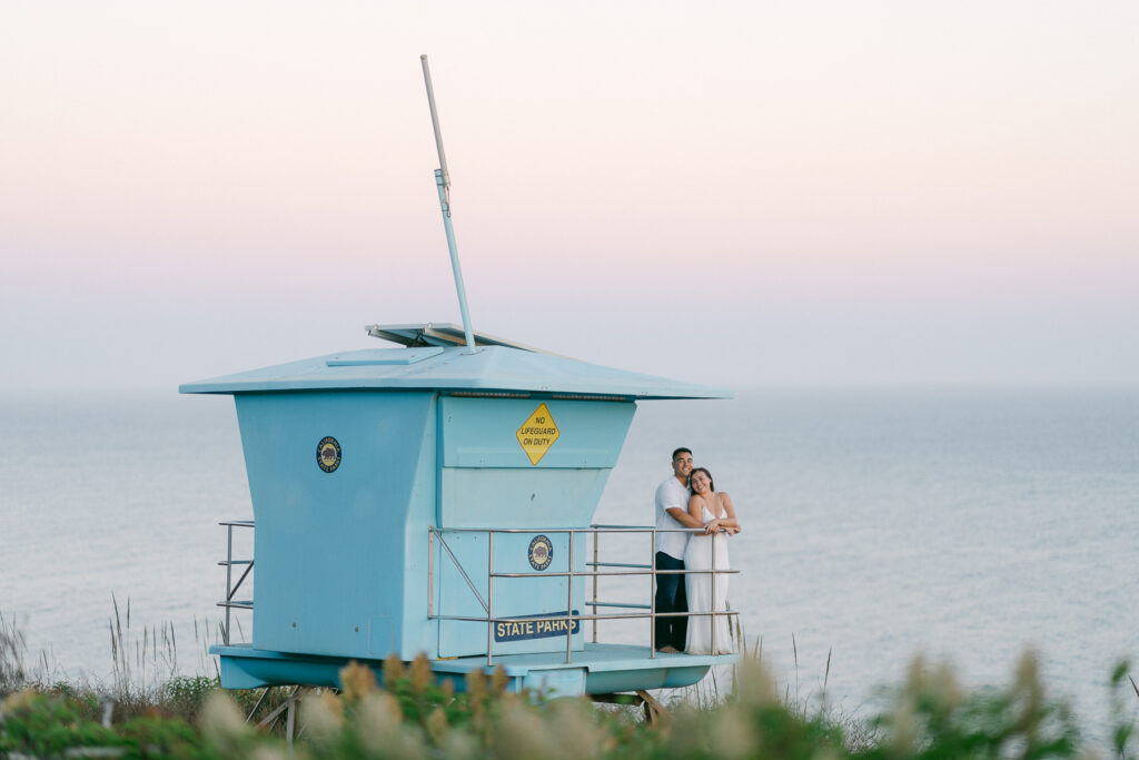 engaged couple standing in the lifeguard tower on a cliff at sunset at el matador beach during their engagement session captured by malibu wedding photographer magnolia west photography