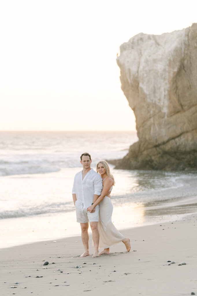 couple along the edge of the water at el matador beach in malibu ca during golden hour girl is wearing a long ivory dress and man is wearing a white shirt and white shorts taken my magnolia west photography best wedding photographer in malibu