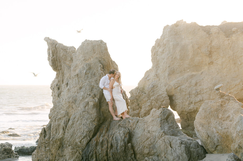 couple standing on the rocks at el matador beach in malibu ca during golden hour girl is wearing a long ivory dress and man is wearing a white shirt and white shorts taken my magnolia west photography best wedding photographer in malibu
