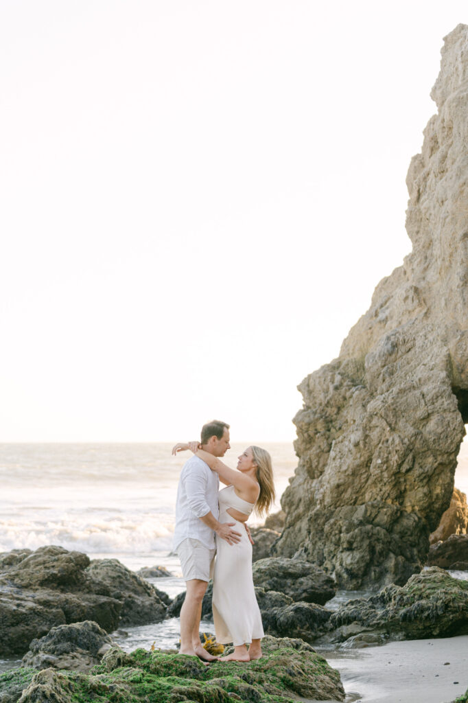 couple standing on the sand in front of the rocks at el matador beach in malibu ca during golden hour girl is wearing a long ivory dress and man is wearing a white shirt and white shorts taken my magnolia west photography best wedding photographer in malibu