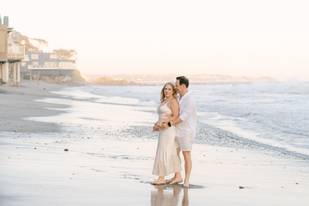 couple along the edge of the water at el matador beach in malibu ca during golden hour girl is wearing a long ivory dress and man is wearing a white shirt and white shorts taken my magnolia west photography best wedding photographer in malibu