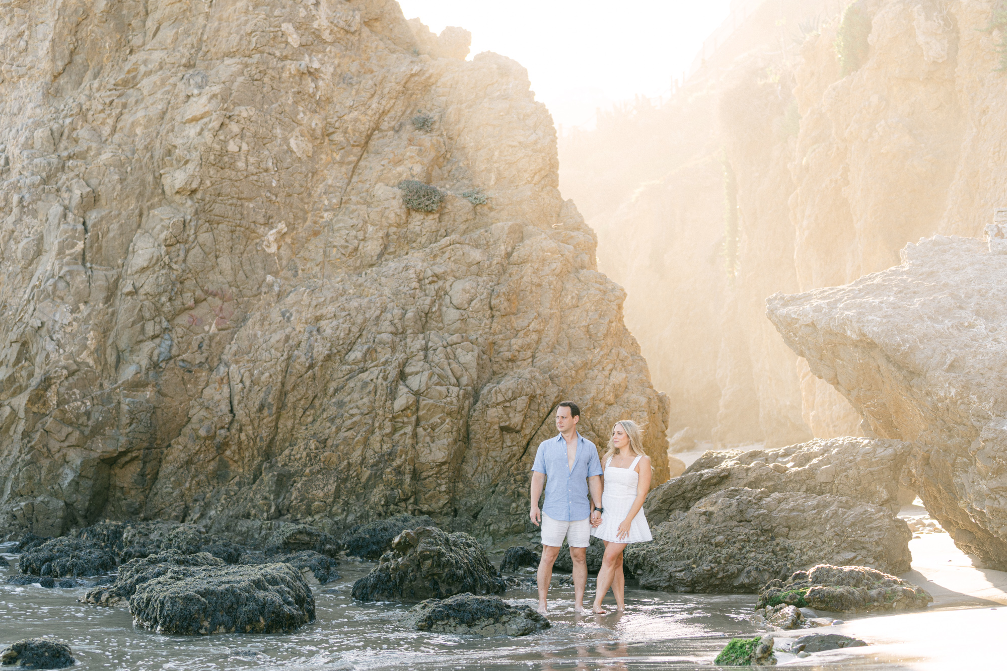 couple standing on the sand in front of the rocks with the light pouring in at el matador beach in malibu ca during golden hour girl is wearing a short white dress and man is wearing a light blue shirt and white shorts taken my magnolia west photography best wedding photographer in malibu