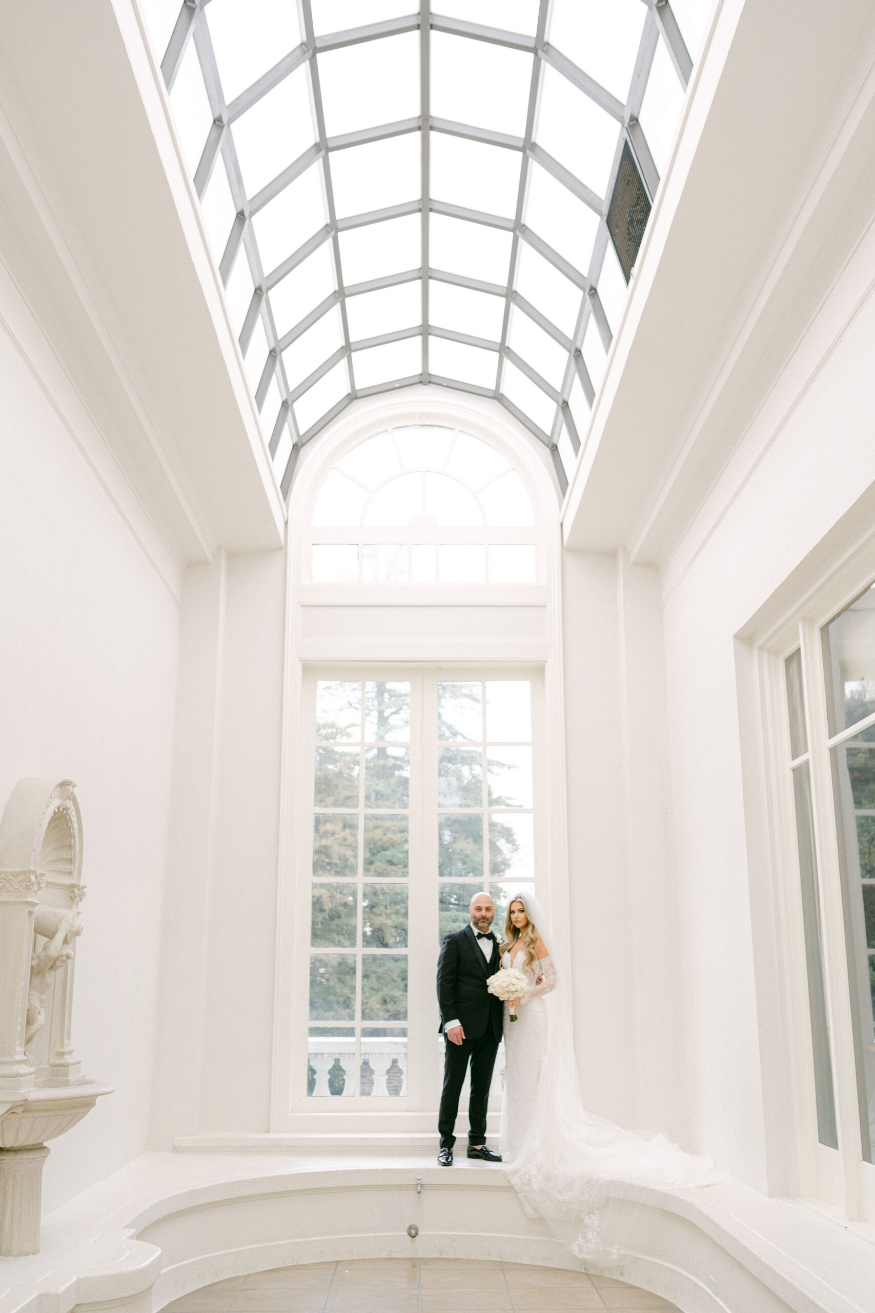 bride and groom staning in a white hall in front of a floor to ceiling window captured by magnolia west photography