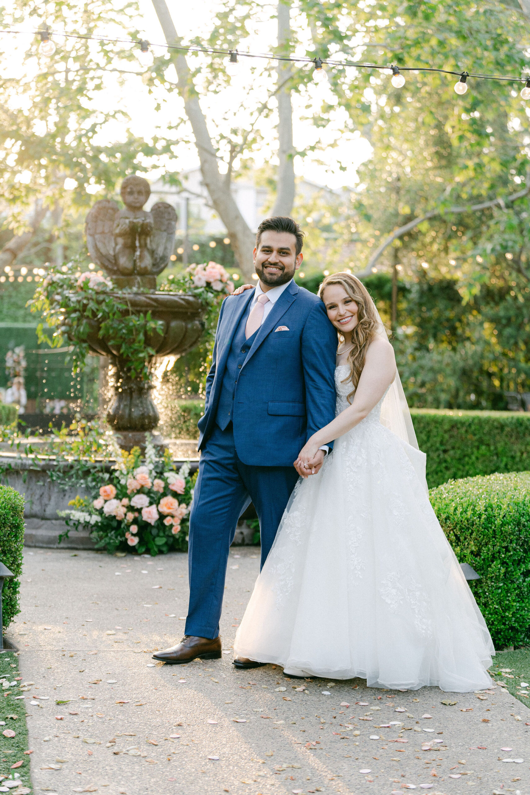 glowy golden hour portrait of an Indian groom in a blue suit with a pink tie and a Jewish bride in a white ballgown posed on the walkway in the garden of The Gardland Hotel in front of the fountain during golden hour